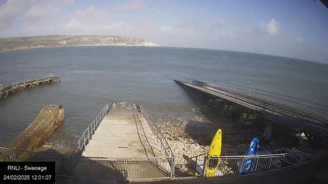 A scenic view of a coastal area featuring a calm sea under a partly cloudy sky. A wooden jetty extends into the water on the left side, with a stone structure near the edge. There is a ramp leading down to the water, surrounded by pebbles and small rocks. Two brightly colored kayaks, one yellow and one blue, are positioned on the right side near a fence. The backdrop includes distant cliffs and greenery, while various small boats can be seen on the water. The timestamp at the bottom indicates the date and time of the image capture.