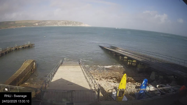 A coastal scene featuring calm water with small waves. In the foreground, there is a wooden ramp leading down to the water, surrounded by a railing. To the right, several brightly colored kayaks in yellow and blue are positioned on the rocky shore. In the background, gentle green hills rise towards a cloudy sky, and a distant coastline is visible across the water. The scene conveys a serene seaside atmosphere.