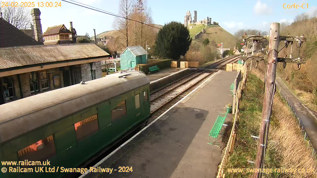 A railway station scene featuring a green vintage train car parked on the platform. The platform is made of stone and has several green benches for seating. In the background, there is a grassy hill crowned with a castle. Clear blue skies are visible, and it appears to be a sunny day. There are trees on either side of the station, and a wooden fence marks the edge of the platform.