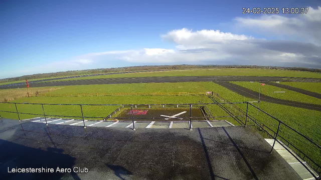 A view from a webcam at Leicestershire Aero Club showing a wide, open landscape. In the foreground, there is a small helipad with a white cross marking in the center. To the left, a railing can be seen, and in the background, there is a green field stretching out toward a gray runway. The sky is blue with some clouds, and a red wind indicator is visible on the left side of the image. The scene is bright and clear, indicating a sunny day.