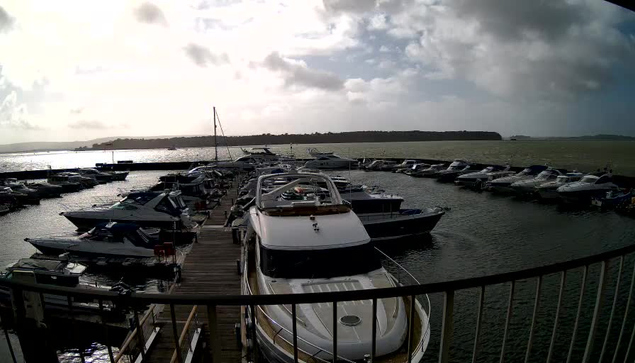 A marina scene featuring several boats docked in the water. In the foreground, a large white yacht with a curved roof is prominently displayed. Surrounding it are smaller boats, some with blue and white colors, all tied to wooden docks. The water reflects the cloudy sky above, and a distant shoreline is visible in the background. The overall atmosphere appears calm and slightly overcast, with gentle waves on the water.