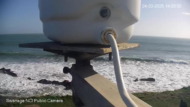 A white container is mounted on a wooden platform, positioned over a rocky cliff with the ocean visible below. The container has a visible valve where a white tube connects to it. The background features waves crashing against rocky outcrops and a clear sky. The date and time are displayed in the upper right corner.