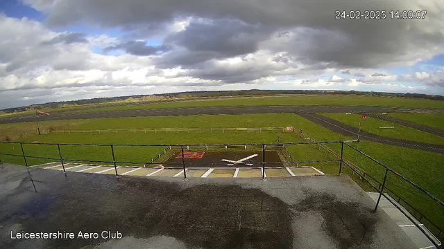 A view from a webcam overlooking an airfield. In the foreground, a gray surface with some moisture is visible. Beyond that, a grassy area extends to a runway, which is marked with white lines. In the center of the image, there is a red square with a white aircraft symbol on it. There are a few scattered clouds in the sky, and the landscape in the background includes fields and distant trees. The time and date are displayed in the top right corner.