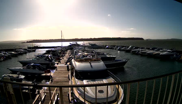 A marina filled with various boats docked along a wooden pier under a bright, sunny sky. The sun is shining in the background, casting a glare on the water's surface. In the foreground, a large white boat is prominent, with several smaller boats visible in the surrounding area. The shoreline in the background is slightly obscured, with trees or land visible. The water appears to be rippling gently.