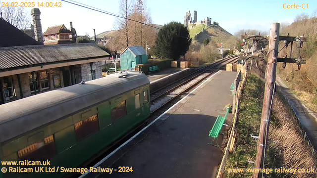 A train is parked at Corfe Castle railway station, featuring a green passenger carriage. The station is surrounded by trees and benches, with a stone building in the background. In the distance, the ruins of Corfe Castle are visible on a hill under a clear blue sky. The tracks are lined with gravel, and the scene is well-lit by sunlight. The image shows a tranquil, rural setting with historic elements.