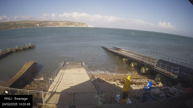 A scenic view of the coast featuring calm blue water stretching out to the horizon under a clear sky with a few clouds. In the foreground, there is a wooden jetty extending into the water, with gentle waves lapping at the structure. To the side, there is a concrete ramp leading down to rocky shore, with some stones visible. A yellow kayak and a blue boat sit on the shore, alongside a railing that surrounds the area. The setting conveys a peaceful seaside atmosphere.