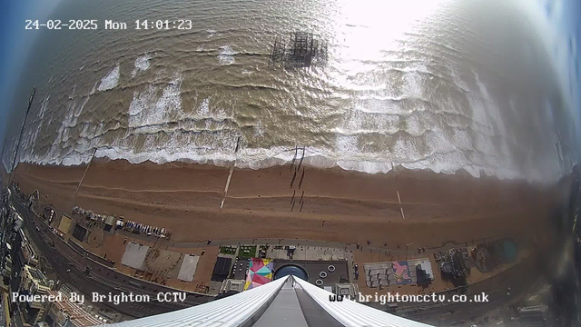 A beach scene captured from above, showing golden sand extending along the bottom of the image. Gentle waves of a light brown ocean ripple towards the shore, creating white foam at the edges. In the background, a structure resembling a pier is partially submerged in the water. Various buildings and a colorful amusement area are visible along the shoreline, with shadows indicating a sunny day. The upper portion of the image is dominated by blue sky, and the date and time are displayed at the top.