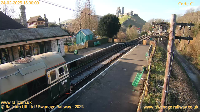 A view of Corfe Castle railway station on a sunny day. In the foreground, a green and cream vintage train is parked on the platform. The station features a stone building with a sloped roof and waiting benches along the platform. To the left, there is a small green shed. On the right, two sets of railway tracks run parallel, leading into the distance. In the background, the iconic ruins of Corfe Castle are positioned on a hill, surrounded by trees. The sky is clear and blue, indicative of a pleasant day.