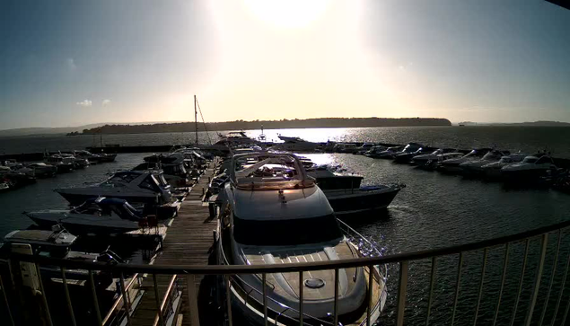 A marina filled with boats is visible under bright sunlight. The foreground shows several motorboats and yachts docked alongside a wooden pier. In the background, more boats are moored, with a shimmering water surface reflecting the sunlight. The sky is mostly clear with a few clouds. A distant landmass is visible on the horizon.