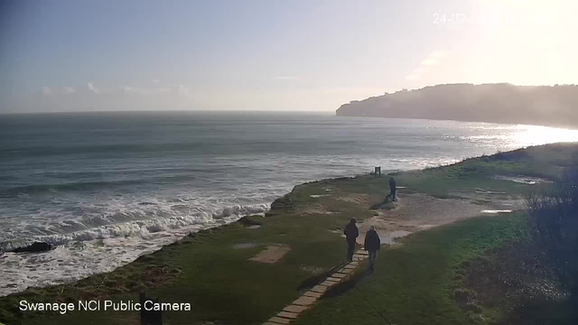 A scenic coastal view featuring two people walking on a grassy path near the water's edge. The ocean is visible with gentle waves lapping at the shore. In the background, there is a cliff under a clear, sunny sky. The light reflects off the water, creating a bright atmosphere. The scene is tranquil and evokes a sense of nature and calmness.