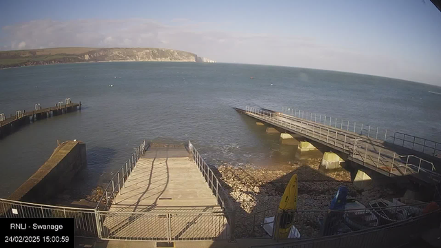 A coastal scene with a view of the sea and a rocky shoreline. In the foreground, there is a concrete ramp leading down to the water, surrounded by a railing. To the left, a jetty extends into the water. In the background, cliffs rise above the water, and the sky is partly cloudy with patches of blue. A yellow kayak is visible on the right side, next to some boats. The date and time are displayed on the bottom left corner.