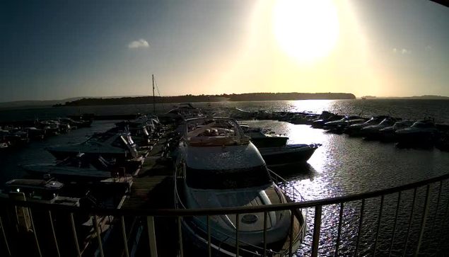 A marina is filled with various boats and yachts moored along a wooden dock. The sun is low in the sky, casting a bright glare over the water, which reflects the sunlight. In the background, there are hills with greenery, while the foreground shows several boats tied to the dock, with some partially out of the water. The scene conveys a calm and serene atmosphere of a daytime waterfront.