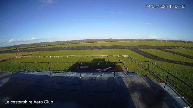 A wide view of an open airfield under a clear blue sky. The foreground shows a railing and a flat surfaced area with a wind direction indicator nearby. In the distance, there is a stretched expanse of green grass leading to a dark asphalt runway, with a few markings and a red sign positioned near its edge. The scene captures a sense of space and tranquility at the Leicestershire Aero Club.