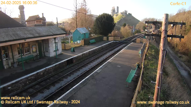 A view of a railway station in Corfe, with stone buildings and green benches along the platform. The station features a roof with a chimney and a small green shed visible in the background. Train tracks lead away from the platform, flanked by trees and hedges. In the distance, a hill with a historic castle can be seen under a clear sky. The image is taken during the day, indicating good visibility.