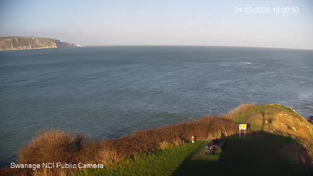 A panoramic view over a calm sea, with gentle waves reflecting sunlight. In the foreground, grass and shrubbery lead to a cliff edge. A single bench is visible near the cliff, and there's a warning sign in the right corner. The sky is mostly clear with a few clouds, and distant cliffs can be seen on the horizon. The image is timestamped in the upper right corner, showing the date and time.