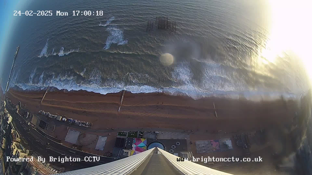 Aerial view of a beach with waves gently breaking on the shore. The sandy beach is partially visible, with some beachgoers. In the distance, a pier extends into the water, where an abandoned structure can be seen. The sky is clear, with sunlight reflecting off the ocean surface. A circular area, possibly a building or attraction, is located on the beach near the bottom of the image. The top of the image shows the edge of a tall building or structure from which the photo is taken.