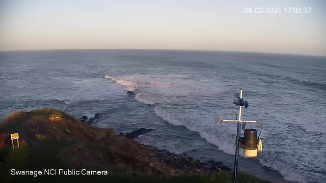 A coastal view with gentle waves lapping against rocky shores. The scene is bathed in soft evening light, with a clear sky above. In the foreground, there is a weather station featuring an anemometer and a cup anemometer mounted on a pole. Nearest to the station, lush green grass and a sign can be seen, warning of the cliff edge, while the horizon meets the calm sea in the distance.