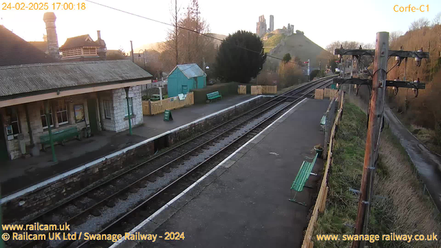 A railway station scene with tracks running through the center. On the left, there is a stone building with a sloped roof and chimney, partially obscured by trees. On the platform, there are several green benches and wooden fencing. To the right, a green shed is visible, alongside more benches. In the background, a hill rises with the ruins of a castle at the top, and the sun is setting, casting a warm glow over the landscape. The sky is clear, and the overall scene has a peaceful, rural atmosphere.