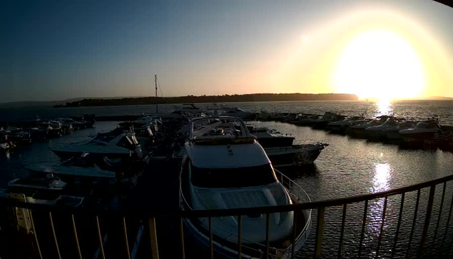 A marina filled with various boats is visible in the foreground, with a bright sunset creating a warm glow over the water. The sky transitions from blue to orange and yellow near the horizon, where the sun is partially hidden. The water reflects the colors of the sky, and small waves can be seen rippling across the surface. In the distance, a shoreline is faintly silhouetted against the sunset. The scene gives a feeling of calm and tranquility.