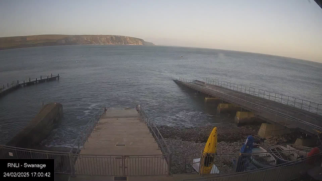 A view of a harbor by the sea during sunset. The foreground features a concrete ramp leading down to the water, with a railing on both sides. To the left, there is a wooden pier extending into the sea. Yellow and blue kayaks are positioned on the shore, and the water appears calm with gentle waves. In the background, cliffs rise along the shoreline under a clear sky, with a few small boats visible on the water's surface.