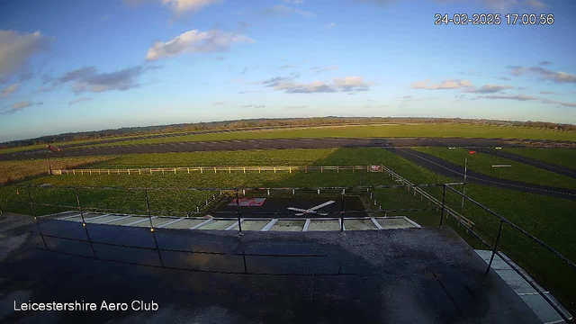 A wide view of a green landscape under a clear blue sky with some scattered clouds. In the foreground, there is a flat surface, likely a platform, which is partially wet. A white "X" marking is visible on the ground, along with a surrounding railing. The background features a runway with asphalt and grass areas, along with a distant horizon lined with trees. The scene conveys a calm day at an airfield, identified as Leicestershire Aero Club.