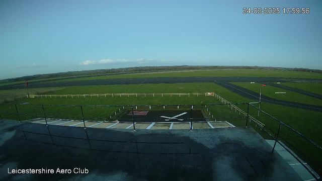 A view from a webcam showing a grassy landscape under a clear blue sky. In the foreground, there is a flat rooftop structure with a railing. Below, a marked area with a white cross symbol is visible, likely indicating a helipad or landing spot. To the left, a red windsock is positioned on a pole, indicating wind direction. The background features a long runway made of dark pavement extending towards the horizon, bordered by a wooden fence and green fields.