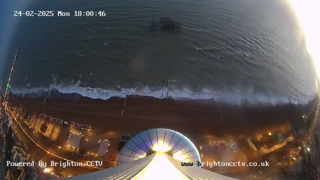 A view from above showing a coastline with a sandy beach meeting turbulent waves. The scene is illuminated by city lights along the beach, and a structure, possibly a pier, can be seen extending into the water. The sky is transitioning from blue to dark as evening approaches. Text at the top indicates the date and time, while a logo at the bottom left signifies the camera's operation.