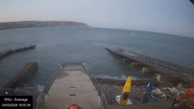 A view of a coastal area featuring a jetty extending into the water. The water is calm with a few boats visible in the distance. On the right side, there are several colorful kayaks stored on the shore. The area is rocky with a ramp leading down to the water. The sky is a light color, indicating early evening.