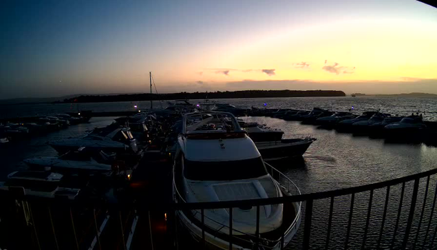 A sunset view over a marina filled with various boats and yachts. The sky displays warm hues of orange and purple as the sun sets on the horizon, casting a soft glow over the water. In the foreground, several boats are moored closely together, with some illuminated by lights. A railing is visible in the foreground, framing the scene. The water appears calm, reflecting the colors of the sky and the silhouettes of the boats.