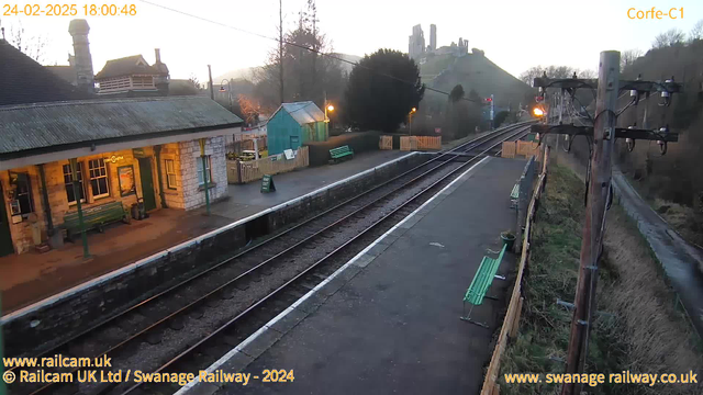 A view of Corfe Castle railway station at dusk. The image shows a stone platform with two green benches on the left side. A traditional-style railway building is partially visible, featuring large windows and a sign that reads "Café Gate." In the background, a hill rises, topped by the ruins of Corfe Castle. The sky is fading from light to dark as evening approaches, and there are faint lights illuminating the station. Railroad tracks run parallel to the platform, leading off into the distance. A power pole with wires can be seen on the right side of the image.