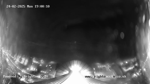 A black and white image captured from a webcam, featuring a view of a city street with blurred lights. The scene is mostly dark with some illuminated areas, suggesting it is night time. There is significant glare in the center, likely from bright lights or reflections. The date and time displayed in the top left corner reads "24-02-2025 Mon 19:00:10." A watermark in the bottom left reads "Powered By Brighton CCTV," and a website link appears at the bottom right.