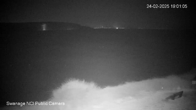 A dark, nighttime scene of a shoreline with waves crashing against rocks in the foreground. The water is mostly calm, reflecting minimal light. In the background, faint lights from the coastline can be seen, indicating a distant town or harbor. The sky is dark, suggesting late evening. A date and time stamp is displayed in the upper right corner, reading "24-02-2025 19:01:05." The camera is identified as "Swanage NCI Public Camera" at the bottom left.