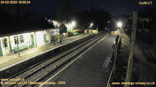 A dimly lit railway station at night, with a stone building featuring large windows and a green bench in front. The platform is empty, with two railway tracks running through the center. In the background, there are wooden fences and some scattered benches. A small shed is visible, illuminated by warm light. The atmosphere is quiet and peaceful, reflecting a sense of stillness.