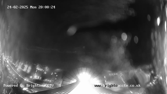 A black and white webcam image shows a brightly lit area in the center with faint outlines of structures and blurred lights extending horizontally on the left side. The date and time displayed at the top indicate February 24, 2025, at 20:00:24. The image appears foggy or misty with a washed-out effect around the bright lights. The text at the bottom left reads "Powered By Brighton CCTV," and there is a web address on the bottom right.