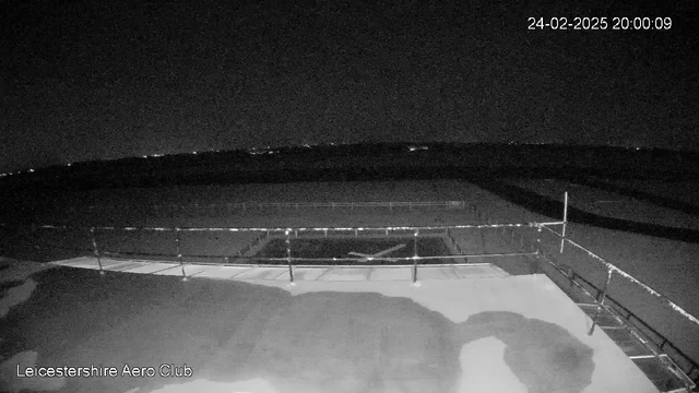 A black and white image showing an open space at night. In the foreground, there is a flat rooftop surface with some limited features and structures. A railing runs along the edge of the roof. The background features a darker sky with distant lights, possibly indicating a horizon or nearby town. The date and time are displayed in the top right corner.