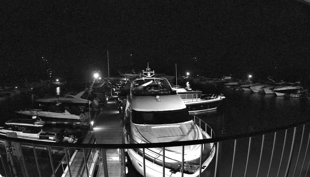 A nighttime image of a marina filled with several boats docked along a wooden pier. The scene is lit by various lights reflecting off the water, creating a peaceful ambiance. Some boats are larger yachts, while others are smaller vessels, all visible in different orientations. The surroundings appear dark, highlighting the illuminated areas. The image conveys a quiet, serene atmosphere typical of a waterfront at night.