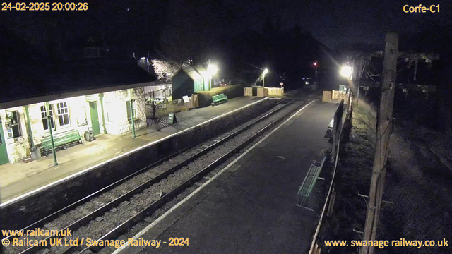 A dimly lit railway station at night, featuring a platform with two railway tracks. To the left, there is a stone building with green accents, including windows and a door. A green bench is visible on the platform. A sign indicating "WAY OUT" is placed upright on the ground. The opposite side of the track has additional seating and a wooden fence. Streetlights illuminate the area, casting soft light, while the background remains dark with trees partially visible.