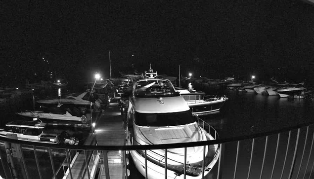 A nighttime scene at a marina, with several boats and yachts docked close to a wooden walkway. The image is in black and white, highlighting the reflections of lights on the water's surface and the silhouettes of the boats. Some boats are illuminated by lights, while others are in shadow. The marina appears peaceful, surrounded by darkness.