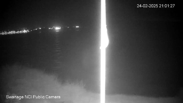 A dimly lit night scene featuring a dark body of water with faint lights visible in the distance, possibly from a shoreline or boats. A vertical white pole is present in the foreground, dominating the left side of the image. The overall atmosphere is dark with low visibility, showcasing a tranquil but obscured coastal view.