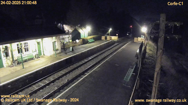 A dimly lit railway station at night, featuring a long platform with two sets of railway tracks running parallel. On the platform, there are benches and a small building with green doors and windows. A wooden fence surrounds part of the station, with a sign indicating "Way Out." The background includes trees and a few distant lights, with the scene mainly illuminated by artificial lighting from the building and lamps along the platform.