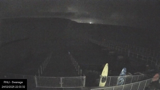 A dark scene captured by a webcam shows a vague outline of a coastal area at night. There are two kayaks in the foreground, one bright yellow and one blue, placed next to a metal fence. The background features faint lights on a distant hillside, indicating a built-up area or town. The image has low visibility with a somber atmosphere due to the nighttime setting.