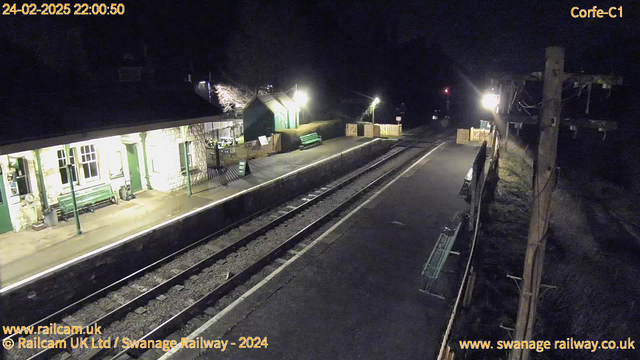 A nighttime view of a railway station platform. The platform is empty, with two railway tracks visible extending into the distance. On the left, there is a stone building with a sloped roof, illuminated by bright lights. A green bench is positioned on the platform. Wooden fencing is visible in the background, alongside another bench and a sign with "WAY OUT" facing the platform. The surrounding area appears dark, with a hint of illumination from nearby lights.