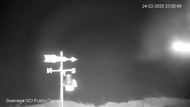 A black and white webcam image showing a weather vane. The vane has arrows indicating north and south. The background is dark with a faint light source on the right side. The bottom portion includes the text "Swanage NCI Public Camera" and a timestamp in the top right corner indicating February 24, 2025, at 11:00 PM.