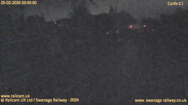 A dark, blurry image captured by a webcam during nighttime. There are faint lights visible in the distance, suggesting buildings or vehicles, with a few red lights that may indicate traffic signals or warning lights. The overall scene is dimly lit, creating a gloomy atmosphere, and the details are not clearly defined due to low visibility. The date and time are displayed in the top left corner.