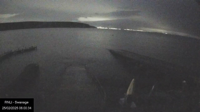 A dimly lit seaside scene captured from a webcam. The water is calm and reflects faint light from the horizon, hinting at a distant shoreline. In the foreground, part of a dock or pier extends into the water. The sky is overcast with clouds, creating a gloomy atmosphere, while faint lights from the shore can be seen peaking through the darkness. The time stamp indicates it's early morning.