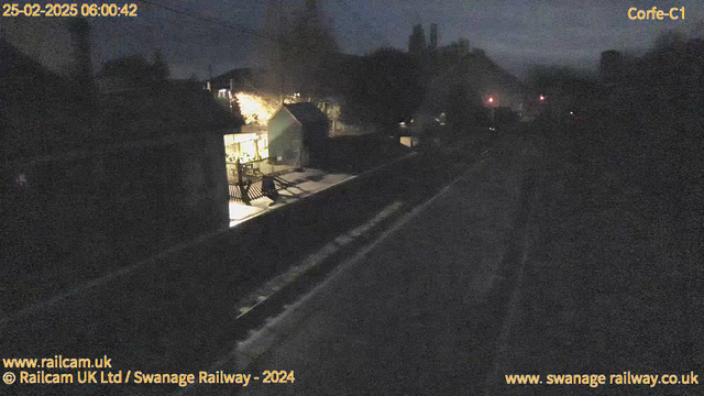 A dimly lit scene at a railway station during early morning. On the left side, a dark structure, possibly a small building or shed, is partially illuminated by lights coming from a nearby area. There are wooden fences and benches visible in front of the building. Train tracks run along the bottom of the image, and the background features silhouettes of trees and possibly a hill. The overall mood is quiet and tranquil, with a few distant lights visible along the tracks.