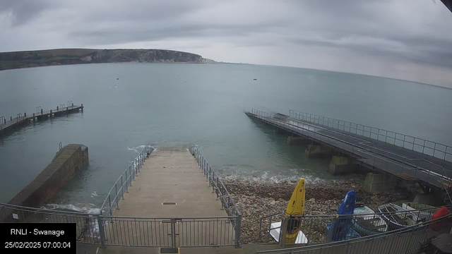 A cloudy scene depicts a calm seaside view. In the foreground, a concrete ramp leads down to the water, with metal railings on either side. To the left, a small, curved wooden dock extends into the water. Various boats are visible on the shore, including a bright yellow kayak and a blue structure. The water appears still, reflecting the overcast sky, with a few distant boats visible on the horizon. The shoreline is rocky, with pebbles scattered along the edge. Overall, the atmosphere feels serene and quiet.