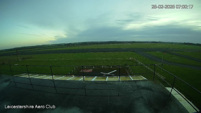 A wide view of an airport scene captured from a webcam. The foreground shows a rooftop railing and a section of the building. Below, there is an airstrip with a small white airplane positioned on a runway. The surrounding area is grassy, and there are some scattered fences along the edges of the runway. The sky appears cloudy with soft light suggesting early morning. The date and time are indicated in the top right corner.