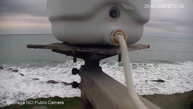 A white cylindrical object is positioned on a metal platform, overlooking a coastal scene. Below, waves crash against rocky shorelines, creating white foam. The sky is overcast, with shades of gray, and the ocean stretches out toward the horizon. The date and time are displayed in the top right corner.