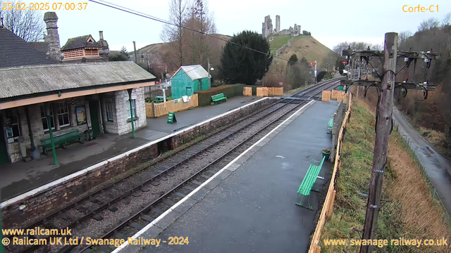 A view of a railway station in Corfe, England, taken from a webcam. The scene shows empty railway tracks with a concrete platform on the left side. Several green benches are placed along the platform. In the background, a hill is visible with a ruined castle at the top. To the right, there are trees and a small wooden building, with a bright green shed nearby. The weather appears cloudy.